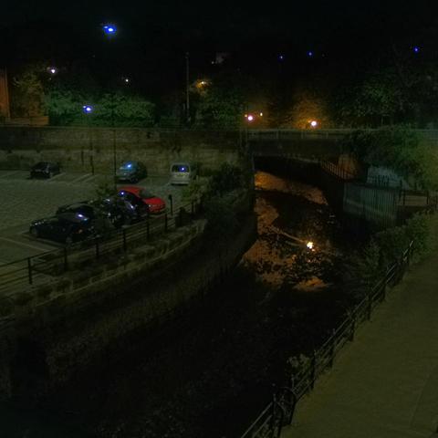 Photograph of the tidal stretch of Ouseburn River at south side of Ouseburn Bridge in Newcastle upon Tyne. To the left (south-west) of the bridge is Cut Bank, and to the right (north-east) is Byker Bank. In the foreground is the pedestrian Riverside Walk and the photo also spans some surrounding built-up areas and trees, with sky in the background. Still image captured from webcam at 03:12 hrs on Monday 5 May 2025.