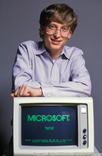 a young white dude with glasses reclining on top of a desktop that has MICROSOFT emblazoned on the screen