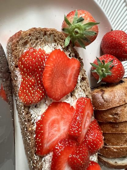 The image features a slice of whole grain bread topped with cream cheese and fresh strawberry slices. Several whole strawberries are placed beside the bread, along with a stack of sliced bread rolls in the background.