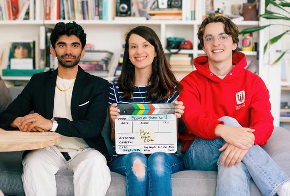 Three people sit on a couch. On the left: a young man with black hair and a beard wearing a black blazer and white pants; in the middle: a woman with long brown hair (me) wearing a blue and white striped t-shirt and blue jeans, holding a clapper with the text: "Fediverse Promo no.1 by Elena Riyen & Sam - May 2nd 2025"; on the right there is a young man with glasses and light brown hair wearing a red hooded sweatshirt. A bookshelf and various items are visible in the background.