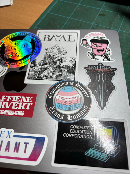 Laptop with a bunch of stickers. A new one with a space marine helmet in trans pride flag colours "transhumans support trans humans"  