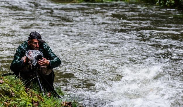 Photographer dressed for rain sitting kneeling down at his tripod, holding the lens in one hand, camera covered in a plastic bag close to a river with water streaming beside him.