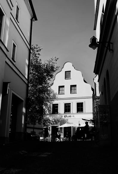 Black and white photo of a white house, illuminated by sunlight at the end of a dark street and the shilouettes of persons in front of the house.