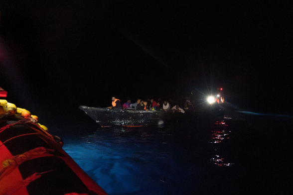 Es ist dunkle Nacht. Von einem RHIB aus Blick zu einem überfüllten, zerschrammt aussehenden Boot. Nur einige der Menschen darin tragen Rettungswesten. Dahinter sind die Scheinwerfer eines weiteren RHIBs zu erkennen.