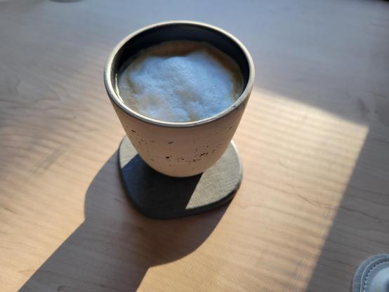 12-ounce, handle-less, off-white ceramic cup with a coffee latte, on a gray diatomaceous-earth coaster on a birch desk. The clay of the cup has many tiny flecks of black and blue. The interior of the cup has a dark-colored glaze. An early-morning sunbeam illuminates a rectangular portion of the desk surface, with the cup casting a shadow.
