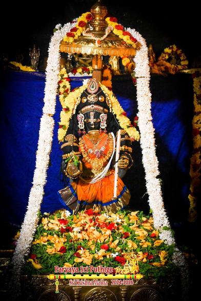 Idol of Hindu deity in peach-orange dress with a blue border. Lots of champaka (yellow) and rose flowers along with holy basil leaves at Krishna's feet. Gold umbrella above Krishna's head, a large white flower garland over the umbrella flowing on either side of Krishna all the way down to the floor. Blue cloth background.