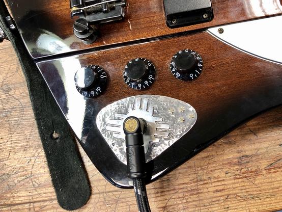 Close-up of a guitar's body. It features knobs for volume and tone, a pickup, and a metallic cover with a textured design. A black and golden vintage style cable is connected to the guitar, resting on a wooden surface.