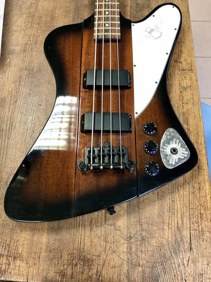 An electric guitar with a unique shape, featuring a dark wood finish, two black pickups, a silver application and a white pickguard. It has four control knobs and is resting on a wooden surface.
