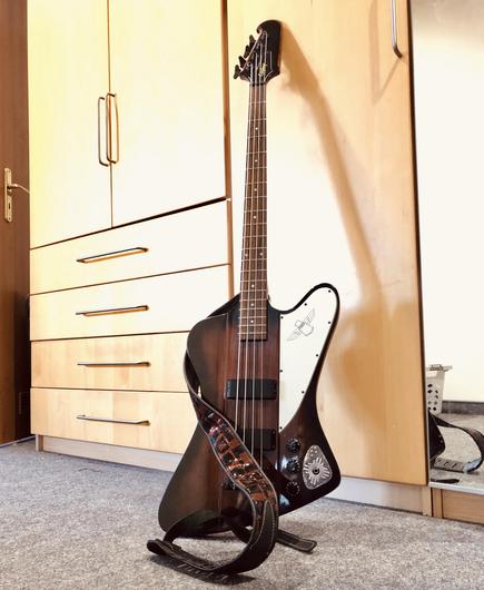 An electric bass guitar with a unique body shape, displayed upright on a carpeted surface. It features a dark wood finish, a white pickguard, a unique silver design and a decorative strap. In the background, there are wooden cabinets and a mirror.