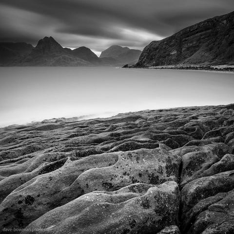 A photograph of the Cuillin mountains viewed from the rocky shoreline at Elgol on the Isle of Skye, where smooth, wave-shaped rocks in the foreground guide the eye toward the jagged peaks in the distance. A long exposure gives the sea a soft, mist-like appearance and stretches the heavy clouds into gentle streaks, adding a calm yet dramatic atmosphere to the scene.