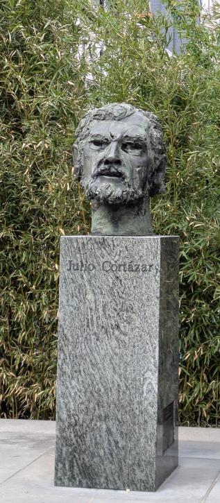 a sculpted head, about 1.5 times lifesize, made from iron in the likeness of Cortázar, on a plinth of grey stone, about 1.70 meters high, with his name in letters of grey metal