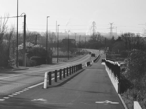 Une piste cyclable bordée d'une route s'étire au loin en noir et blanc. Des pylônes de ligne à haute tension sont visibles en arrière plan