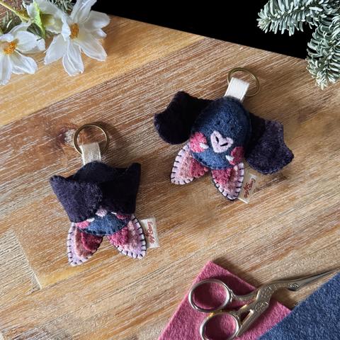 Two bat felt keychains photographed on a black and wooden background, surrounded with decorative plants and felt. 
The bats have a dark grey, egg-shaped body with a dark pink blush, a small heart-shaped, light pink eyes, and two big ears, the inside of them is a three-toned pink. 
One of the bat has it’s wings open, and the other one has them closed over it’s nose, it’s eyes peeking out. Two magnets are hidden in the tips of the wings, allowing them to close. 
