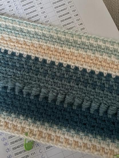 Part of the beginning of a crocheted blanket on a piece of paper with data. The colour of the row represent the maximum temperaturen of the day. 