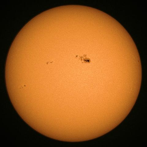 A close-up image of the Sun, showing its bright orange surface with several sunspots visible as dark patches.