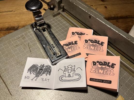 a stack of cute 2 inch square zines sits on a paper cutter next to a stapler