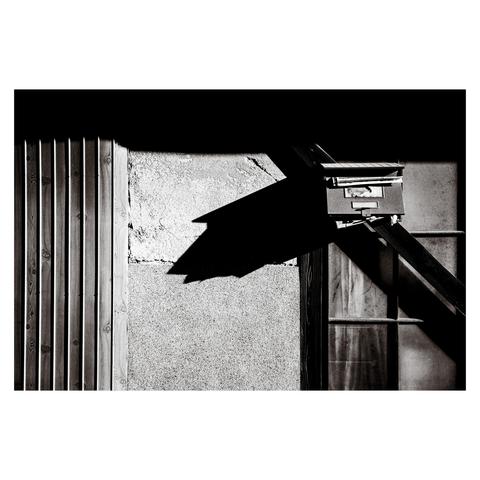 A weathered mailbox juts from a traditional exterior wall, its overstuffed contents creating a dramatic, exaggerated shadow in the harsh afternoon light. The surrounding textures—wood paneling, rough plaster, and frosted glass—form a collage of surfaces slowly surrendering to time.