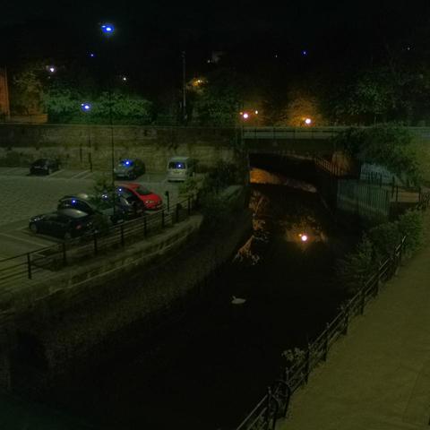 Photograph of the tidal stretch of Ouseburn River at south side of Ouseburn Bridge in Newcastle upon Tyne. To the left (south-west) of the bridge is Cut Bank, and to the right (north-east) is Byker Bank. In the foreground is the pedestrian Riverside Walk and the photo also spans some surrounding built-up areas and trees, with sky in the background. Still image captured from webcam at 03:12 hrs on Tuesday 6 May 2025.