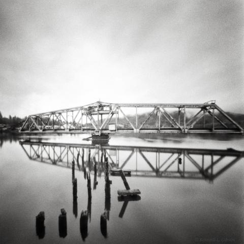 Black and white photo of trestle on river with pilings in the foreground.