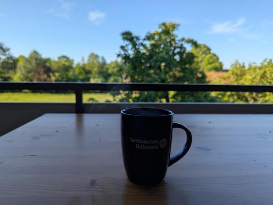 Kaffeetasse mit Aufschrift "Technisches Hilfswerk" auf einem Tisch auf einem Balkon vor der Baumkrone eines Apfelbaumes und blauem Himmel.