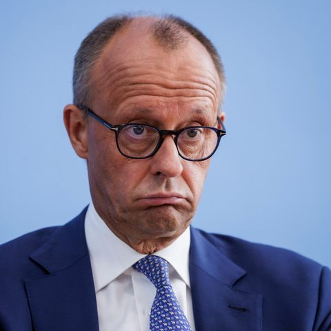 Das Bild zeigt Friedrich Merz im Anzug vor einem hellblauen Hintergrund. Er traegt eine runde Brille mit dunklem Rahmen, hat einen kurzen, licht gewordenen Haarkranz mit deutlich sichtbarer Stirnglatze. Seine Gesichtszuege wirken angespannt oder leicht verstoert, mit nach unten gezogenen Mundwinkeln und gerunzelter Stirn – ein Ausdruck, der an ein skeptisches oder unzufriedenes Stirnrunzeln erinnert. Er traegt ein dunkelblaues Sakko, ein weisses Hemd und eine hellblaue Krawatte mit einem dezenten Muster. Das Bild ist scharf, gut ausgeleuchtet und auf Brusthoehe aufgenommen, wodurch das Gesicht und die Mimik stark im Mittelpunkt stehen.