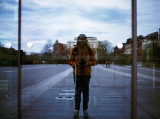 A person is standing outside in an open, urban square, facing a reflective glass surface and taking a photo of their reflection. The individual is wearing a burnt orange jacket, jeans, and sneakers, with a backpack and a beanie. Their camera is held at waist level. The reflection creates a layered effect with the cityscape in the background, including buildings, bare trees, and a cloudy sky. The overall mood is calm and introspective, with cool, slightly muted colors.