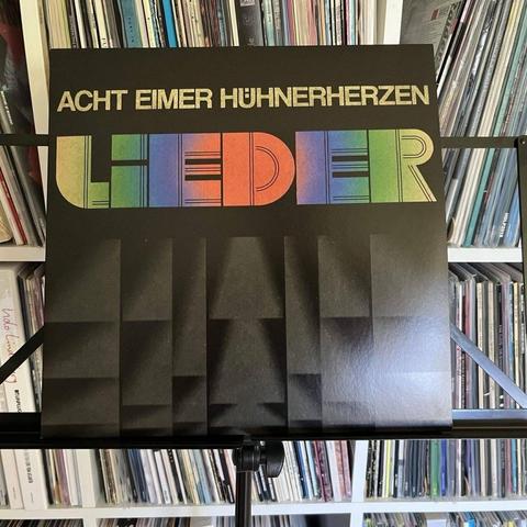 Ein Albumcover mit dem Titel "Acht Eimer Hühnherzen - Lieder" mit fetter, farbiger Typografie auf schwarzem Hintergrund. Das Album ist auf einem Ständer ausgestellt, mit einer Sammlung von Schallplatten im Hintergrund.