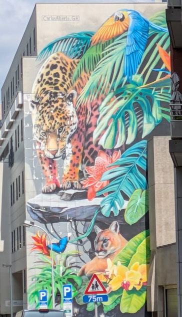 a colorful about five-story mural depicting a leopard, a puma, a grand parrot, and large, sheltering leaves