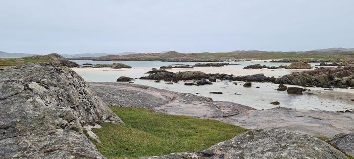 Another bay, this time with white sand and clear blue sky, despite the grey sky above. The beach is speckled with large rocks, and the rocks & sand are slick from the retreating tide.
