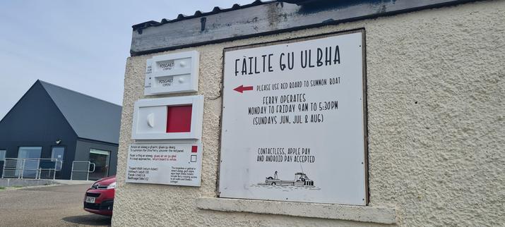 Signage on the side of the building, with three white wooden sliders that can move back and forth. The biggest one is for the ferry, and as explained on the panel below in English and Gaelic, “to summon the ferry, uncover the red panel. As the boat approaches, return board to white.” The two smaller sliders show whether businesses on the island are open or closed - both are open today. A larger sign next to the sliders explains the ferry runs Monday to Friday, 9am-5:30pm, and on Sundays in June, July and August, and that you can pay by contactless, Apple Pay, and Android Pay.