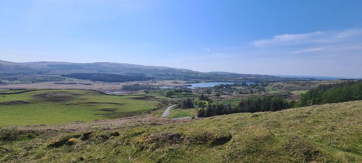 A view from a hill top overlooking a glen. A river widens into a small bay, with some houses on one side. The image though is mostly filled with green hills, fields and small wooded areas.