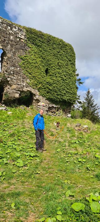 A man - grey curly hair, blue jacket - standing with a small brown and white dog in front of the ruins of a castle. Vegetation has consumed much of the castle’s remaining wall.