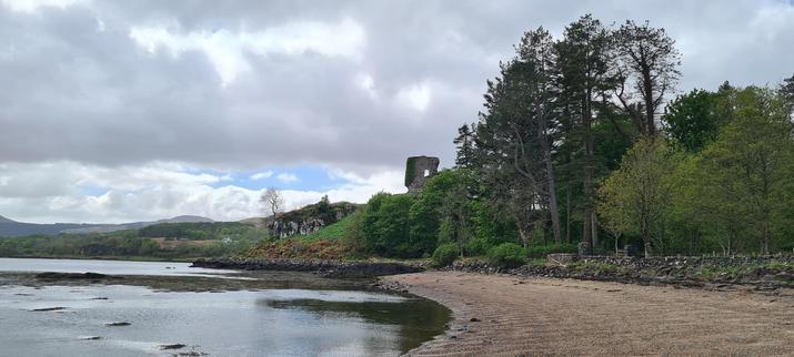 The ruins of the same castle from the previous photo, but this time at more of a distance. The castle rests on a rocky outcrop, over a curved sandy-ish bay.