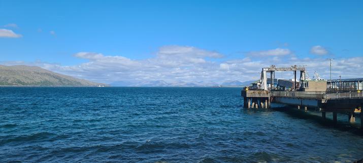 A small ferry terminal stretches out into a calm blue sea. Hazy hills are visible in the distance.