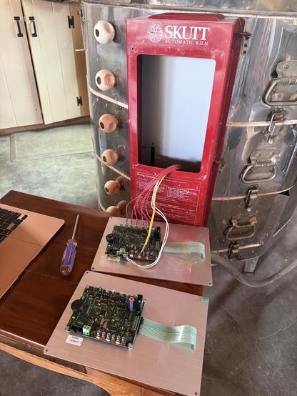 A Skutt electric kiln is visible, alongside two control panels placed on a wooden table. A screwdriver is nearby, and there are wires connecting to the kiln. A concrete floor and wooden cabinets can be seen in the background 