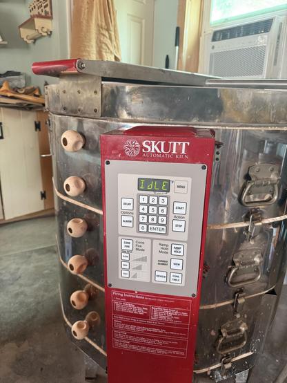 Post-replacement photo: An electric kiln is shown in a workshop setting. The kiln features a digital control panel with an "Idle" status display. Several small clay pieces are positioned on the kiln's exterior. The background includes various craft supplies and a window.