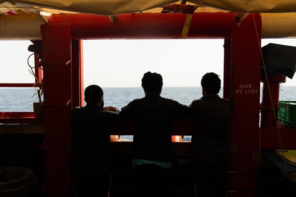 Drei Personen stehen an einem Fenster der Ocean Viking und blicken auf das offene Meer hinaus.