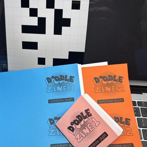 a process pic of a finished issue 1 of a pixel doodle zine and uncut sheets for issues 2 and 3 underneath. a computer screen is behind with a few chunky black pixels colored in over a white canvas.
