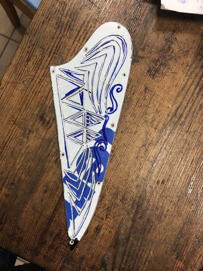 An intricately designed piece, featuring geometric patterns and blue outlines on a white plastic foil cover on a metal sheet background. The lines are engraved in the metal sheet underneath the cover, which will be removed soon. It is placed on a wooden surface. 