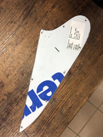 A fragment of a white and blue sign or panel rests on a wooden surface, featuring text in blue. The panel has several holes along its edge, possibly for mounting, and a label with printed information attached to it.