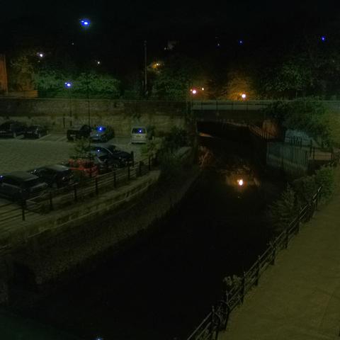 Photograph of the tidal stretch of Ouseburn River at south side of Ouseburn Bridge in Newcastle upon Tyne. To the left (south-west) of the bridge is Cut Bank, and to the right (north-east) is Byker Bank. In the foreground is the pedestrian Riverside Walk and the photo also spans some surrounding built-up areas and trees, with sky in the background. Still image captured from webcam at 03:12 hrs on Wednesday 7 May 2025.