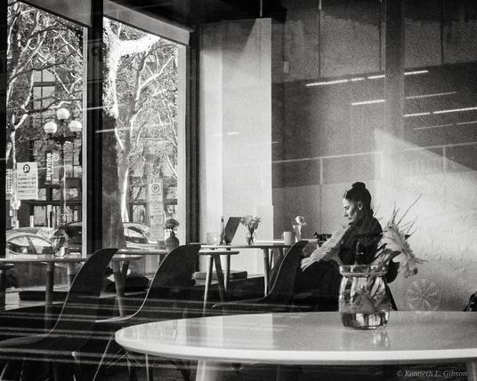 Black and white photograph of a lone woman in a cage surrounded by empty tables and chairs.  A table with flowers in a jar in the foreground.