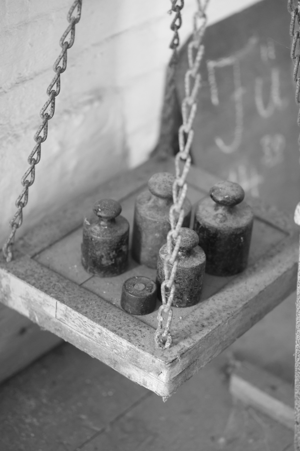 Schwarz-/Weiß-Fotografie zeigt 5 Eichgewichte auf einer Holzplatte, die an 4 Ketten hängt. Gesehen in einer alten Mühle zum Abwiegen von Getreide- und Mehlsäcken. 