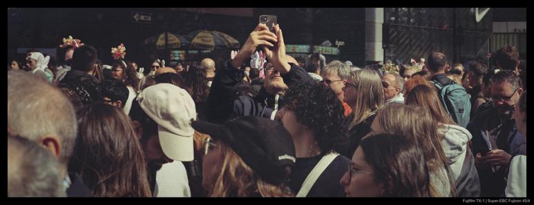 A person lifts their phone above a crowd in order to take a picture to the left of the prevailing direction of movement.