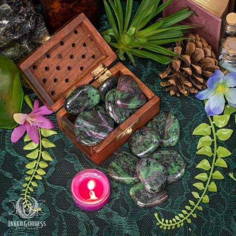 Six specimens of the "Ruby Zoisite Slice" product are arranged on a dark lace-covered surface. Several more specimens fill a wooden box with a hinged lid. They are in mottled shades of green from deep forest to bright grass, with white and red inclusions. They are flat, tumbled smooth, and polished. Presented by Inked Goddess Creations.