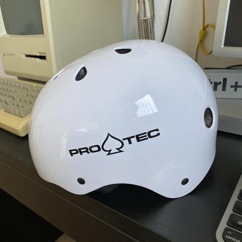 Picture of a glossy white protective helmet for skating, seen from the side. It has the “Pro-Tec” logo printed in black on it.