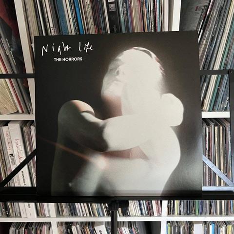 Ein Vinyl-Schallplattencover mit dem Titel "Night Life" von The Horrors, das ein stilisiertes Bild einer Person mit Unschärfeeffekt vor einem Hintergrund von Regalen mit verschiedenen Schallplatten zeigt.