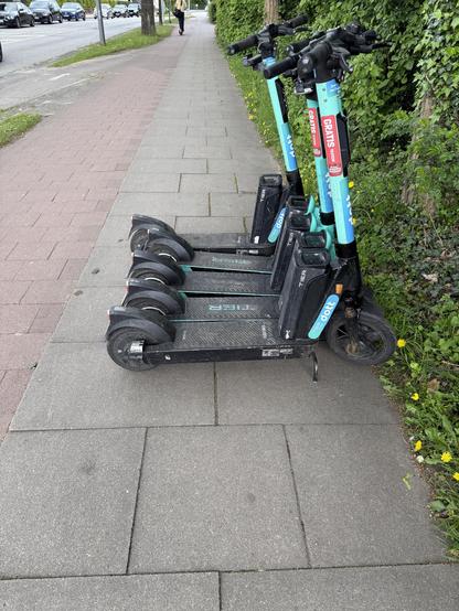 Vier dicht nebeneinander quer auf dem Gehweg abgestellte E-Scooter der Firma DOTT. Die Roller versperren den Fußweg bis auf ca 10 cm. 
