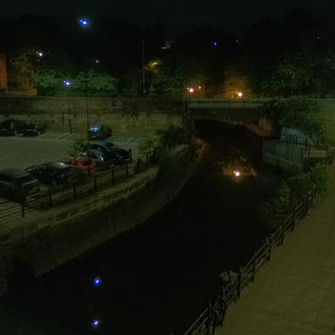 Photograph of the tidal stretch of Ouseburn River at south side of Ouseburn Bridge in Newcastle upon Tyne. To the left (south-west) of the bridge is Cut Bank, and to the right (north-east) is Byker Bank. In the foreground is the pedestrian Riverside Walk and the photo also spans some surrounding built-up areas and trees, with sky in the background. Still image captured from webcam at 03:12 hrs on Thursday 8 May 2025.