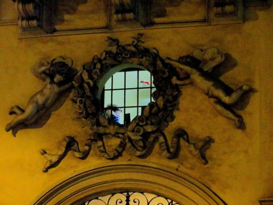A blurry night image of a round window over a passageway. The window is framed by a stucco flower crown, held by two puttis. Inside the window, a lightly lit and ordinary-looking room is visible, with a person sitting at a desk.